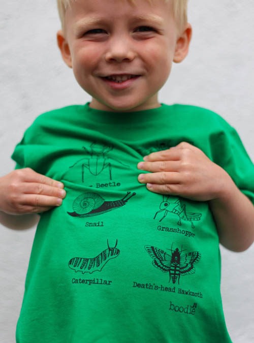 Green organic T-shirt featuring 6 bugs. Kid model wearing the T-shirt with his hands on chest