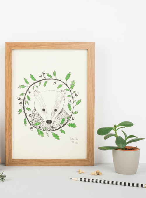 Badger and oak art print. Photo of a framed print featuring an illustration of a badger surrounded by an oak wreath. Green and black print on a natural white background.