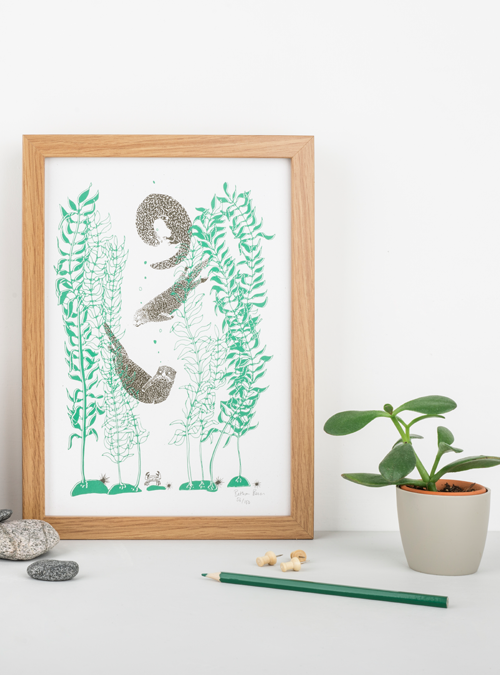 Photo of a art print featuring 3 otters playing in kelp. Screen printed by hand, the otters are brown and the kelp green. Photo is of a framed print with props such as plants and pencils surrounding it