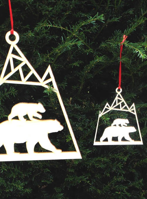 2 wooden decorations hanging in the woods. Polar bear wooden decorations one large and one smaller both with a mum and baby bear under a mountain. Laser cut onto birch ply and hanging on a red ribbon