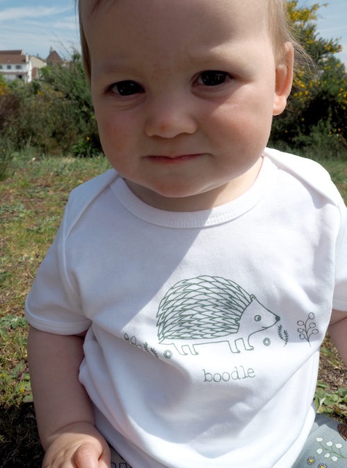 Baby wearing white organic T-shirt featuring an illustration of a hedgehog with foliage