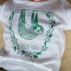 Photo of a baby wearing a sleepy sloth organic white T-shirt. Illustration of a sloth hanging from a branch