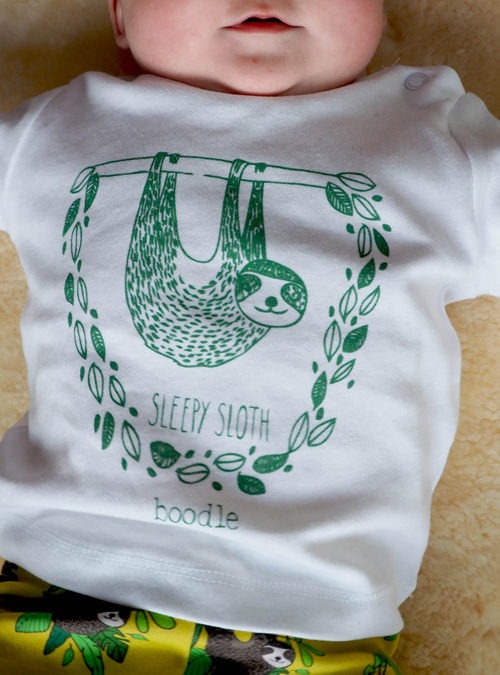 Photo of a baby wearing a sleepy sloth organic white T-shirt. Illustration of a sloth hanging from a branch