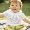 Baby wearing Organic baby Sloth baby t-shirt. White T-shirt with a green print of a sloth hanging from a branch with the text 'Sleepy sloth' underneath