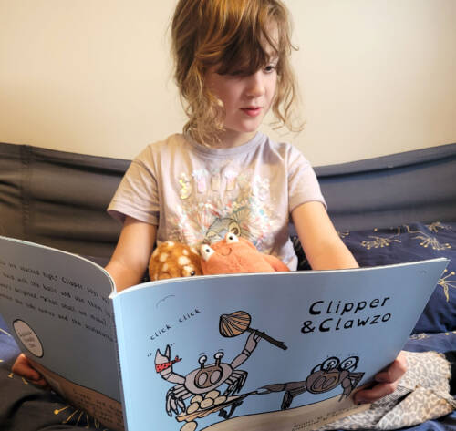 A child model reading the childrens book Clipper and Clawzo sitting on a bed with her cuddly toys