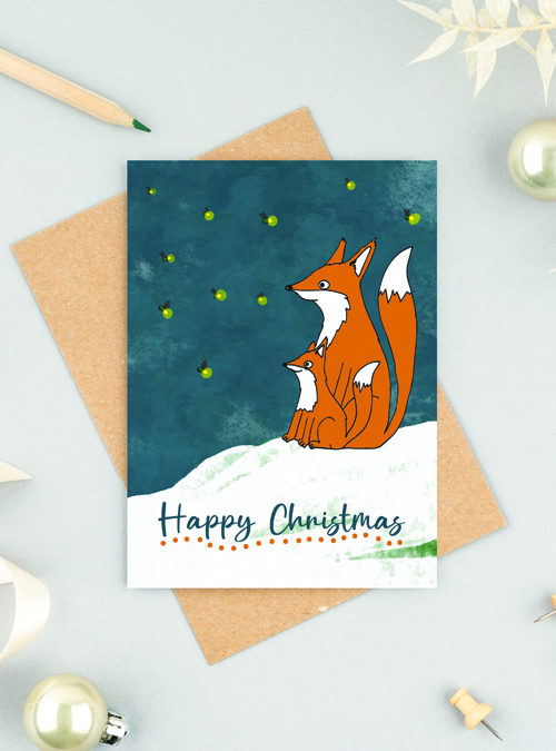 Fox and firefly Christmas card sitting on a brown envelope with festive props